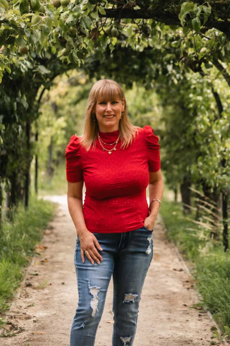 323A3663 Levensgenieter vrouw in rode blouse op tuinpad met groen bladerdak.